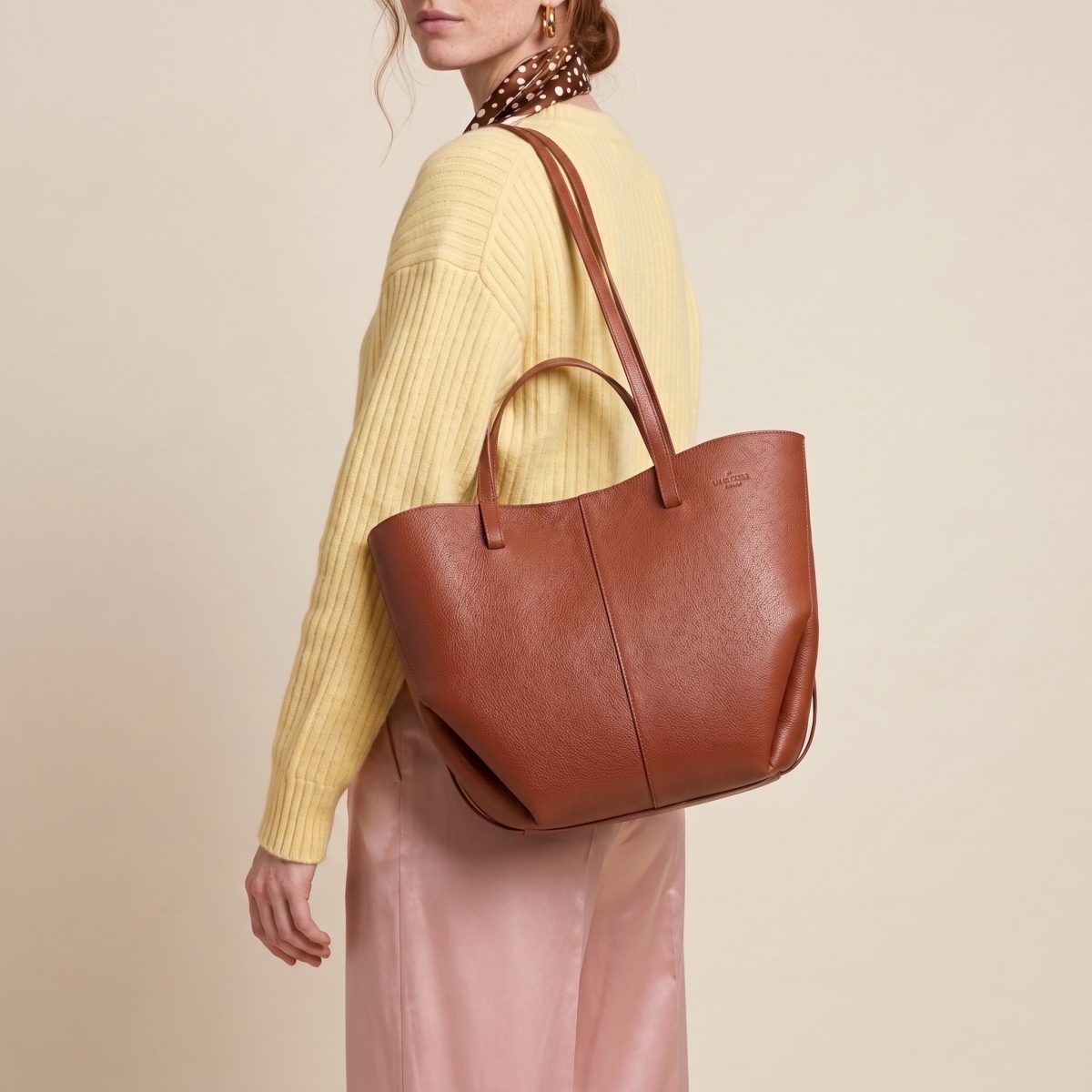 La tote in pelle cognac di Via Verdi portata sulla spalla, con un maglione a coste giallo burro, su fondo color sabbia