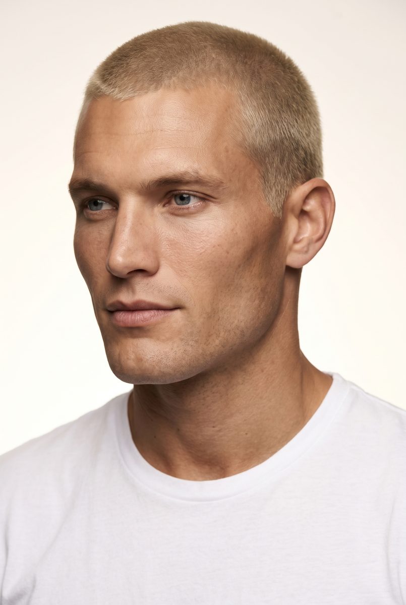 Masc, candidato del cast: ritratto di tre quarti, capelli biondi rasati, t-shirt bianca, fondo chiaro