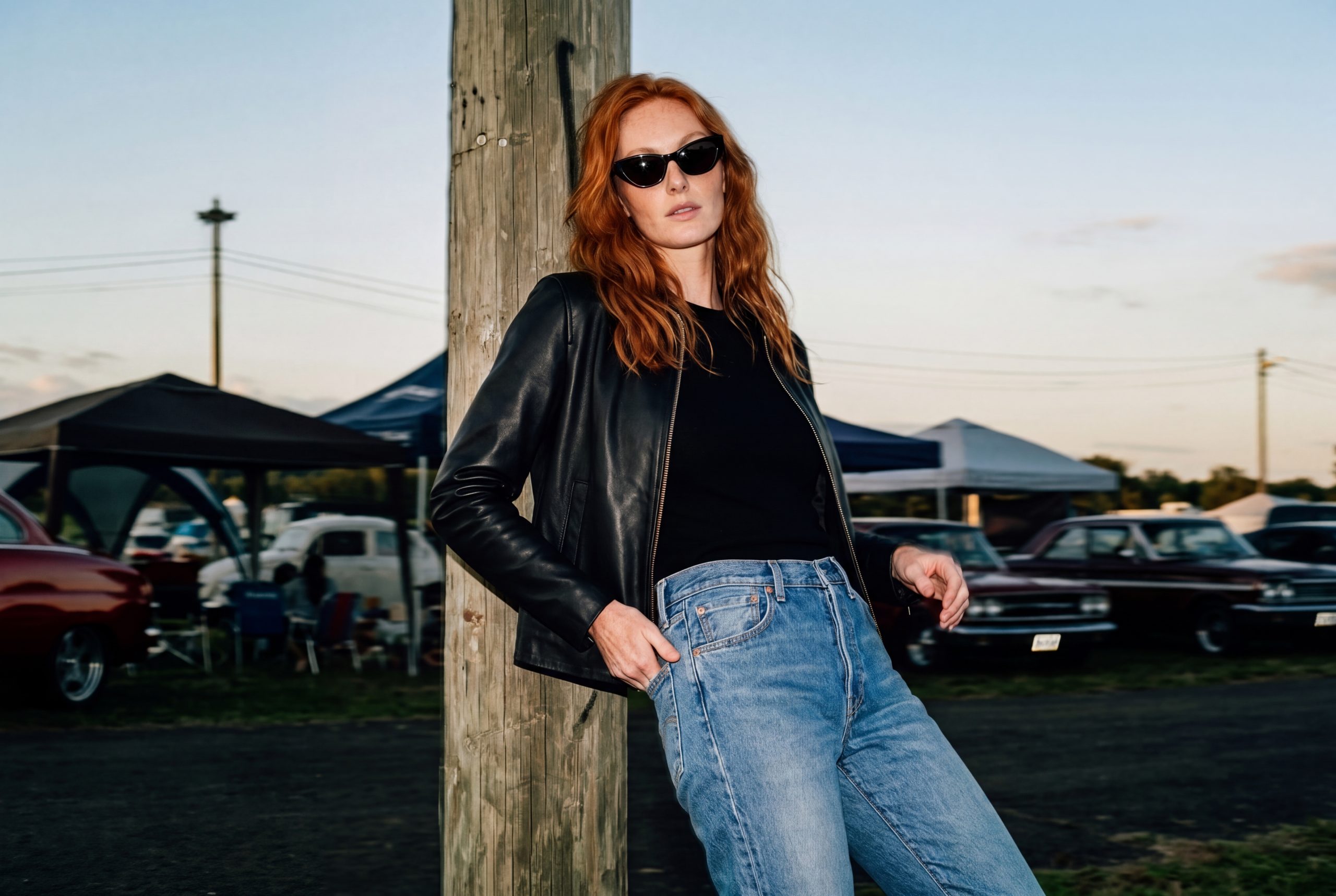 Elisabeth in giubbotto di pelle e jeans appoggiata a un palo, a un raduno di auto d’epoca al tramonto
